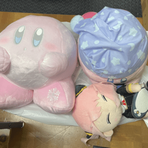 Three oversized plush toys on a wooden floor: a large pink round character, a pastel purple star-patterned plush, and a pink-haired cartoon figure in a black outfit.