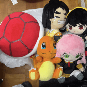 A group of plush toys on the floor: a large red segmented plush, Charmander, a pink-haired girl, and two anime characters.