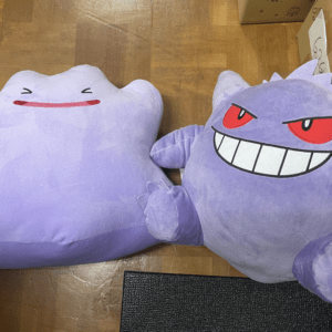 Two purple plush toys lying on a wooden floor: a cloud-shaped one with a simple red smile and closed eyes, and a Gengar-like creature with red eyes and a wide toothy grin nearby near cardboard boxes.
