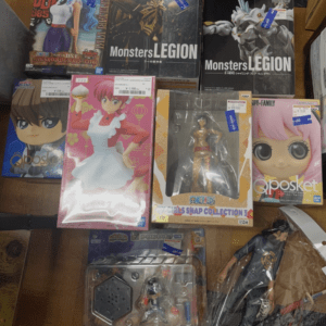 Assorted anime and video game figure boxes on a wooden table, including Qposket and Monsters Legion sets with various characters awaiting display.