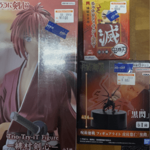 Three boxed anime figures on a wooden surface: a large box with a pink-haired character in red attire on the left, a red box with bold Japanese text and a small figure on top in the middle, and a dark box with a figure on a black pedestal in the bottom-right.
