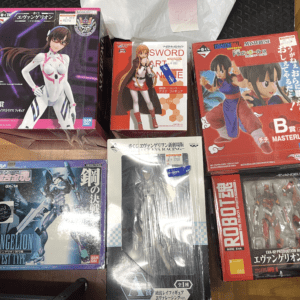 Collection of Evangelion model kits and figures in colorful boxes on a wooden floor, including EVA-01/02/mecha figures and Masterline boxes.