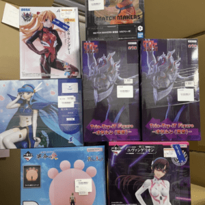 Stack of anime figure boxes on a brown background, featuring characters from Dragon Ball, Evangelion, and other series with visible brand logos like Bandai and SEGA.