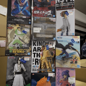 Stack of boxed anime figures on a store display, featuring Dragon Ball, One Piece, and other character boxes stacked on shelves in a storefront.