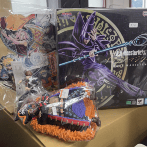 Boxed anime merchandise featuring a large purple Dark Magician figure with a blue staff, alongside illustrated character cutouts on the left and a plush toy with orange accents in plastic wrapping in front.