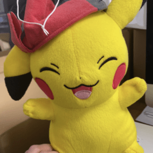 Yellow Pikachu plush wearing a red and gray hat with a white string, smiling with rosy cheeks.