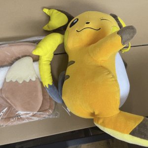 Yellow Pikachu plush toy leaning on a brown backpack in a plastic bag on a cardboard box surface.  Articulos Japan (Post Box Japan)