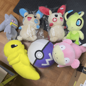 Assorted plush toys on a wooden floor, including a yellow worm, pink creature, green alien-like toy, two beige bunny plushies with colorful ears, a gray cat, and a large Poké Ball stuffed toy.