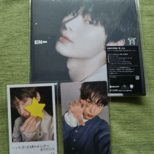 CD album with a close-up portrait on the cover; in front are two photo cards, one with a yellow star obscuring the face and the other showing the person posing with a hand gesture