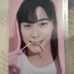Portrait of a smiling young man with black hair wearing a pink hoodie, biting a pink string in his mouth, on a photo card.