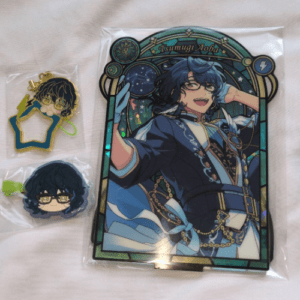 Stained-glass style card of a blue-haired anime character in a blue outfit labeled 'Tsumugi Aoba', in plastic packaging, beside two small chibi keychains with glasses and accessories.