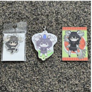 Three chibi anime character keychains/cards laid on a carpet: left a horned character in a plastic bag, middle a horned charm with a blue orb on its head, and right a horned girl in black on a green-orange card.