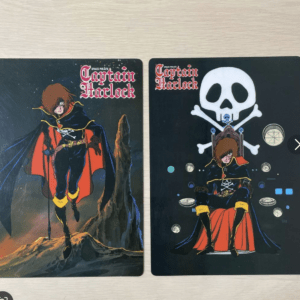 Two Captain Harlock posters on a light wooden surface: left shows the hero standing on rocks with cape flowing, right shows him seated under a skull-and-crossbones motif with space icons in the background.