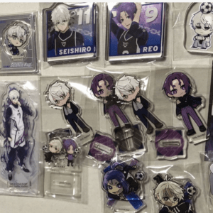 Assorted anime merchandise laid out on a white surface: chibi and full-body acrylic standees, badges, keychains, and buttons featuring white-haired and purple-haired characters.