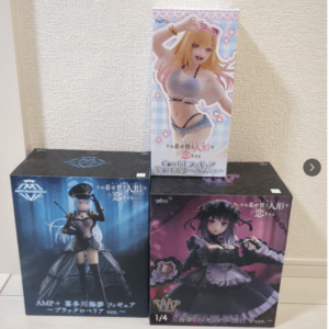 Three anime figure boxes on the floor: a blue box left, a purple box right, and a pink box on top featuring a blonde girl in a bikini.