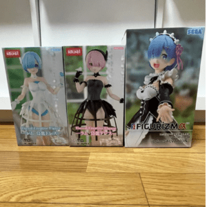 Three boxed collectible figures on a wooden floor: blue-haired figure in white dress, pink-haired in black dress, and blue-haired maid in black-and-white outfit.