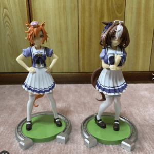 Two anime-style female figurines in matching purple-and-white sailor uniforms stand on green circular bases, posing with hands on hips.
