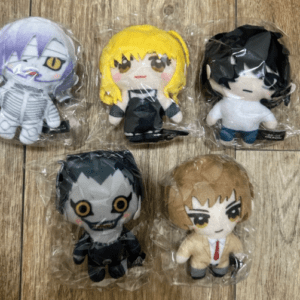 Five small plush figurines in clear plastic bags arranged on a wooden floor: top row left a purple-haired character in a white outfit, center a yellow-haired character in black, right a dark-haired character in a white shirt and blue pants; bottom row a gray, yellow-eyed creature and a tan-haired character in a beige suit with a red tie.
