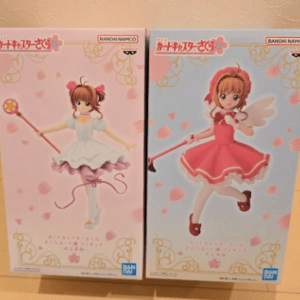 Two boxed anime figures of winged magical girls on a wooden surface: left figure in a light pink dress with a wand, right figure in a pink/red dress with wings and wand, pink and blue packaging with decorative gold swirls.