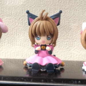Three cute chibi figurines in pink and red dresses sit on a glossy black shelf, one with pink ears, one with black cat ears, and one with a red bow.