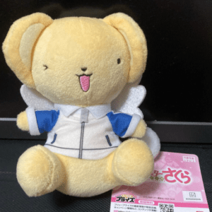 Yellow plush bear wearing a white jacket with blue sleeves and small wings, sitting on a surface with a pink tag nearby.