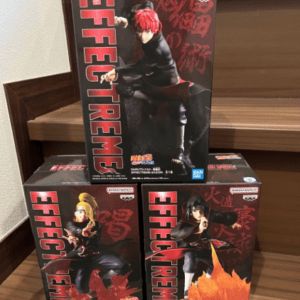 Three boxed anime action figures arranged on a wooden staircase, red-and-black packaging with illustrated characters and Bandai Namco logos.