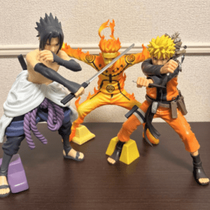 Three anime action figures posed in a dramatic battle stance on a shelf: a dark-haired fighter with a sword on the left, a flaming orange-clad figure center, and a blond-haired fighter on the right.
