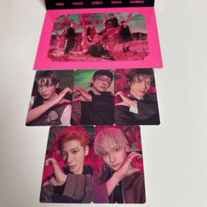 K-pop photo card set on a white surface with a neon pink folder above; multiple glossy cards show group members posing and forming heart shapes with their hands.
