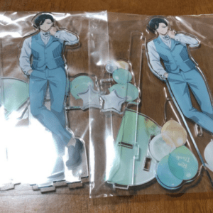 Two acrylic standee cutouts of a man in a light blue suit inside clear plastic packaging, with stand bases and circular connector pieces nearby.