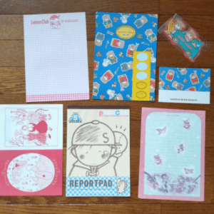 Assorted colorful stationery items on a wooden floor: letter paper, envelopes, postcards, and stickers.