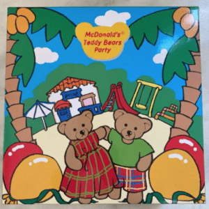 Two teddy bears in plaid outfits stand together at a sunny playground with palm trees and a slide, on a McDonald's Teddy Bears Party cover.