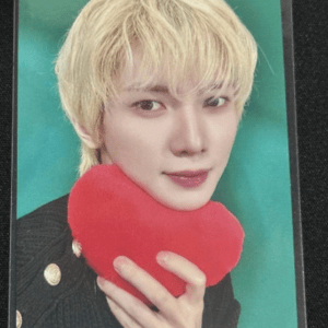 Person with blond hair holding a red heart-shaped plush against a green background.