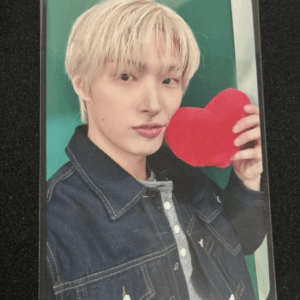 Young person with blond hair posing and holding a red heart-shaped prop, wearing a dark denim jacket against a green background.
