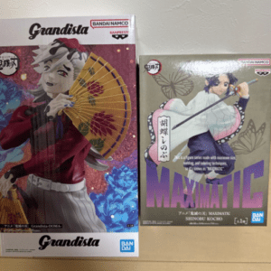 Two boxed anime figurines on a floor: a large Grandista figure with a red outfit and yellow parasol on the left, and a Maximatic figure with a sword and pink umbrella on the right. Both boxes show Bandai Namco branding.