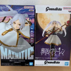 Two Bandai Namco action figure boxes on a table: left MAXIMATIC edition with a white-haired armored character; right Grandista edition showing the same character in a white dress with a staff.