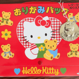 Red Hello Kitty letter-set box featuring a central Hello Kitty illustration with bear characters, a hanging silver charm, and Japanese text at the top; bottom reads 'Hello Kitty' with blue and green hearts.