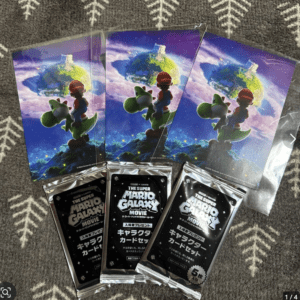 Posters featuring Mario and Yoshi standing on a rocky globe in space, with a bright purple sky and distant stars behind them (The Super Mario Galaxy Movie). The three posters lay on a patterned background, along with promotional card packs labeled in Japanese for The Super Mario Galaxy Movie.