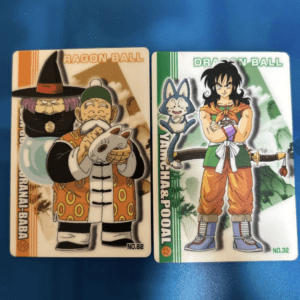 Two Dragon Ball trading cards on a blue surface; left card shows an old man in a robe with a cat, right card shows a muscular hero in orange pants with a sword and a green background.