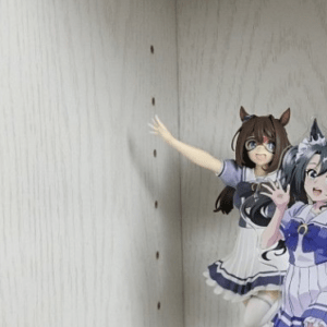 Shelf display of anime-style figurines inside a light-colored cabinet, with multiple characters arranged along the bottom edge and one figure reaching toward the back wall.