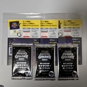 Three black sealed packs labeled The Super Mario Galaxy Movie, arranged in front of yellow QR promo sheets behind them.