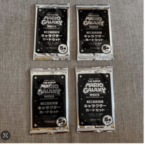 Four sealed 'The Super Mario Galaxy Movie' character card set packets on a textured gray surface, each with a black starry design and '5枚入り' label.