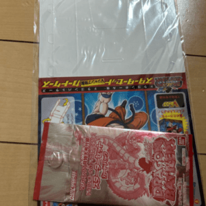 White handheld device in plastic wrap atop a wooden floor, partially covered by a red sealed packet over a colorful Japanese game cover.