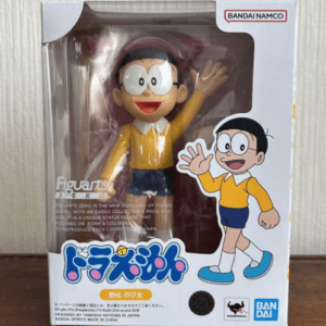 Doraemon figure Nobita inside clear packaging, wearing a yellow shirt and blue shorts, waving; box shows cartoon Nobita illustration and Bandai Namco logo.