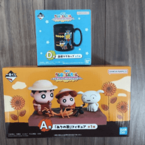 Two Bandai collectible figure boxes on a wooden surface: a blue mug box sits above an orange sunflower-themed box featuring three characters and a white cat.