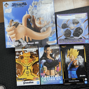Boxed Bandai anime figures on display: a large One Piece Luffy figure in the top left with a giant raised fist, plus other characters in colorful boxes (Naruto/DBZ-style lineups) in the lower row; packaging shows branding like Grandista and Glitter & Glamours.  Articulos Japan (Post Box Japan)
