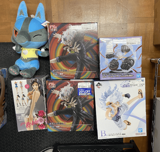 Captura De Pantalla 2026-05-04 a Las 170329 - Articulos Japan (post Box Japan) Blue plush cat sits on a wooden floor beside assorted anime figure boxes. Articulos Japan (Post Box Japan)