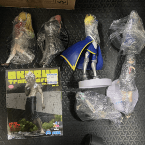 Several action figures wrapped in plastic are laid out on a dark patterned floor, with a boxed figure featuring a muscular character nearby and a small blue cape displayed on a stand.  Articulos Japan (Post Box Japan)