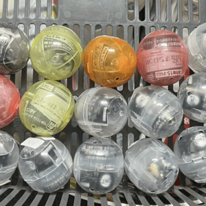 Top-down view of a gray basket filled with assorted translucent toy capsules in colors pink, yellow, orange, red, green, and clear. Each capsule likely contains a small surprise item (gashapon).