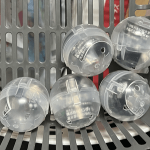 Five clear plastic capsules resting in a gray vending rack, each containing small items inside.