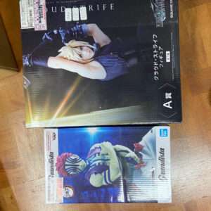 Two boxed action figures sit on a wooden surface: a large black box featuring Cloud Strife, and a smaller white/blue box with a colorful striped character.